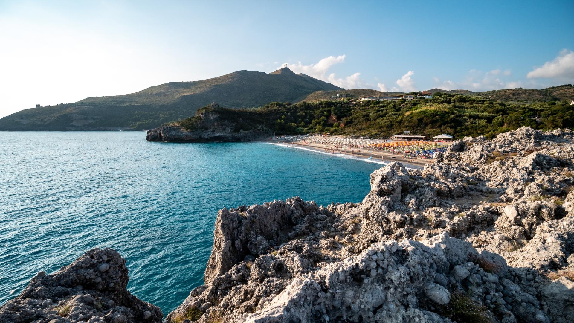 Spiaggia con ombrelloni colorati, mare azzurro e scogliere in una baia pittoresca.